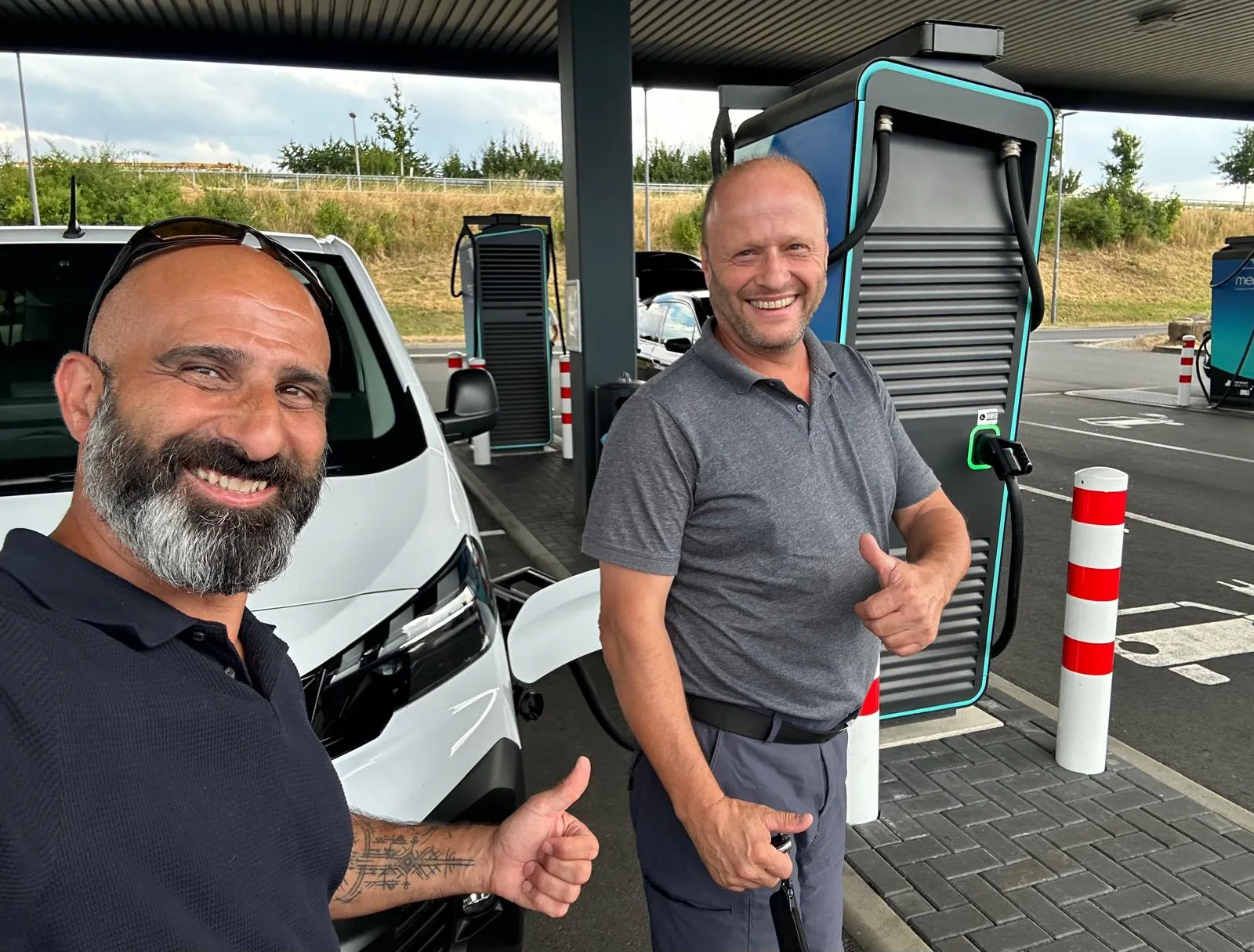 Nader Samadi Tehrani und der Erste Beigeordnete Peter Gute stehen an einer Schnellladesäule neben dem weißen Bürgerbus und heben beide den Daumen