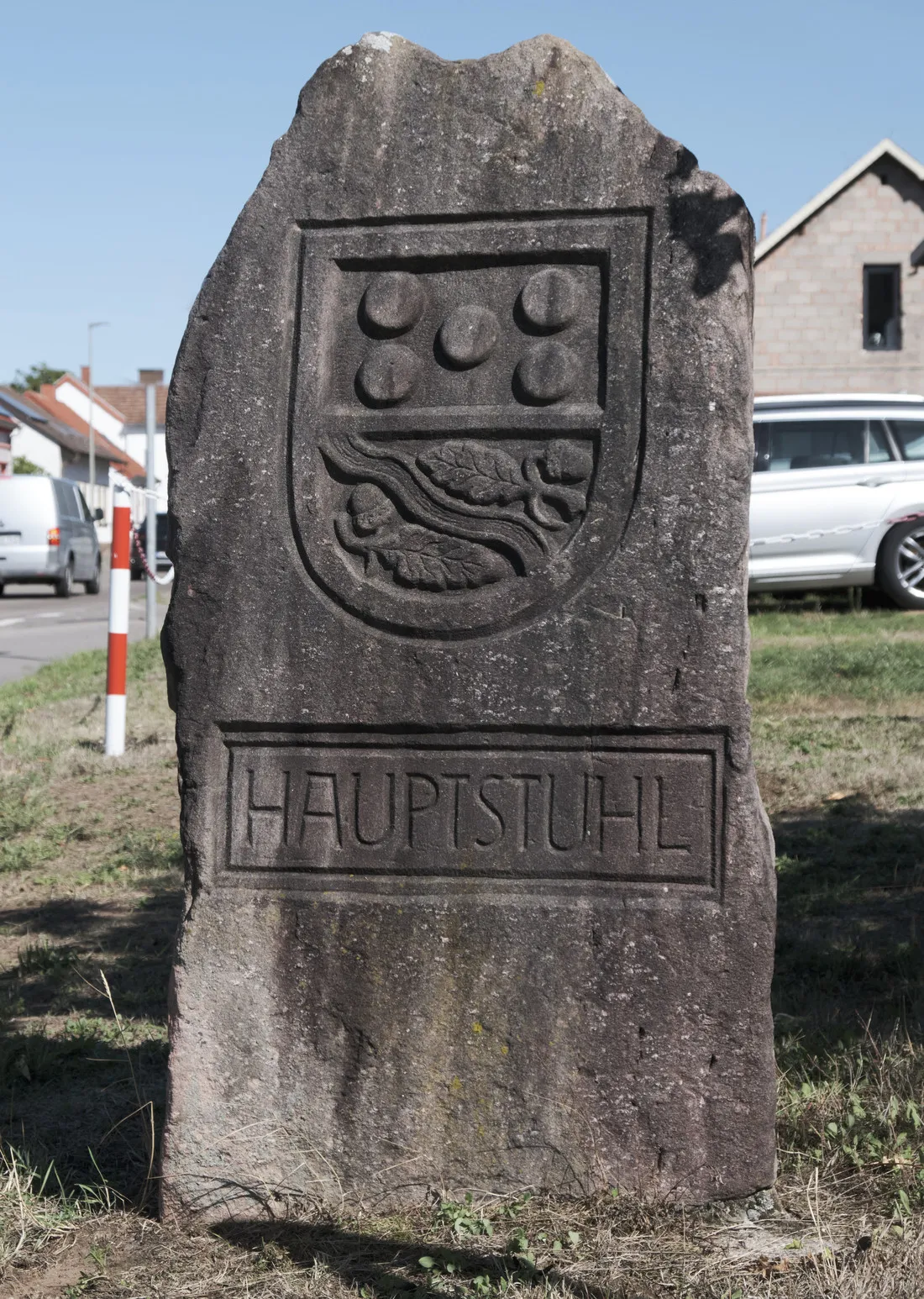Der Ortsstein von Hauptstuhl auf der Wiese am Ortseingang: ein mannshoher Sandstein mit dem eingehauenen Gemeindewappen und dem Schriftzug HAUPTSTUHL, dahinter die Häuser des Dorfes.