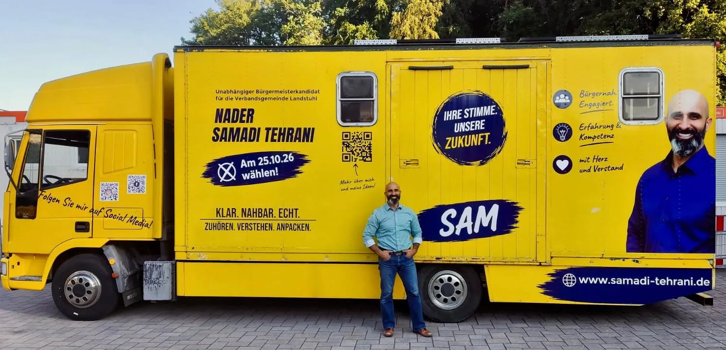 Der gelbe Tourlastwagen in voller Länge, davor steht Nader Samadi Tehrani. Auf der Fahrzeugseite stehen sein Name, der Satz „Ihre Stimme. Unsere Zukunft.“ und der Hinweis, am 25.10.26 zu wählen.